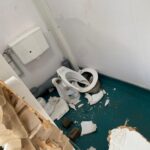 Damaged bathroom with a broken toilet seat and scattered debris on a teal floor across the corner of the room.