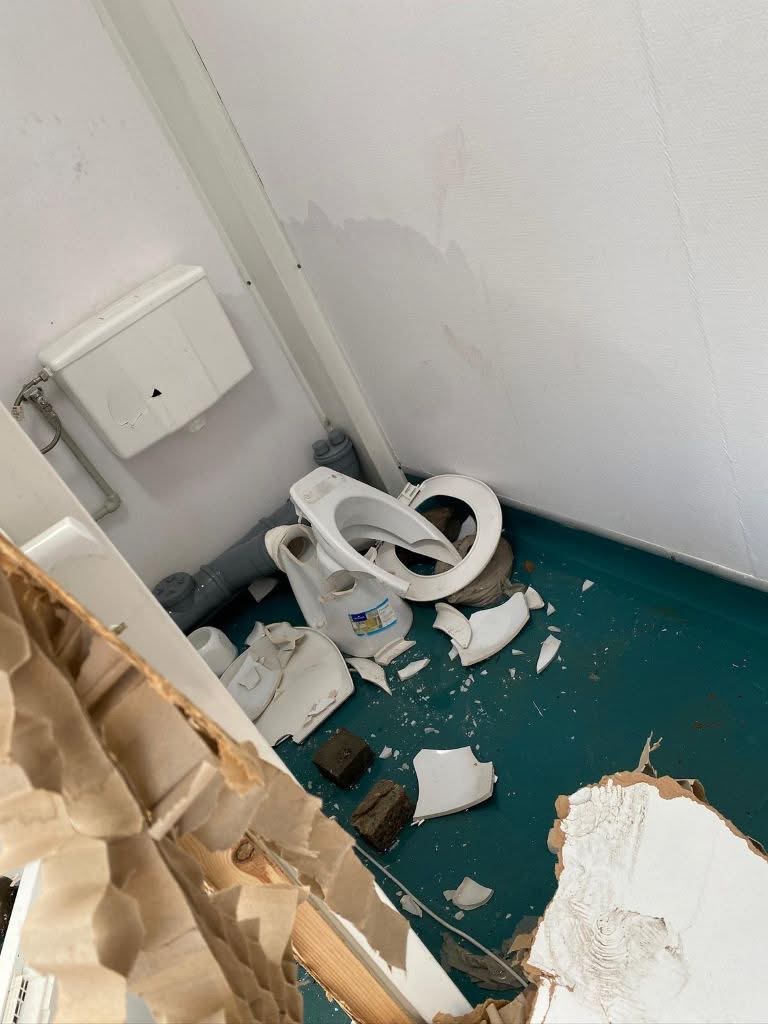 Damaged bathroom with a broken toilet seat and scattered debris on a teal floor across the corner of the room.