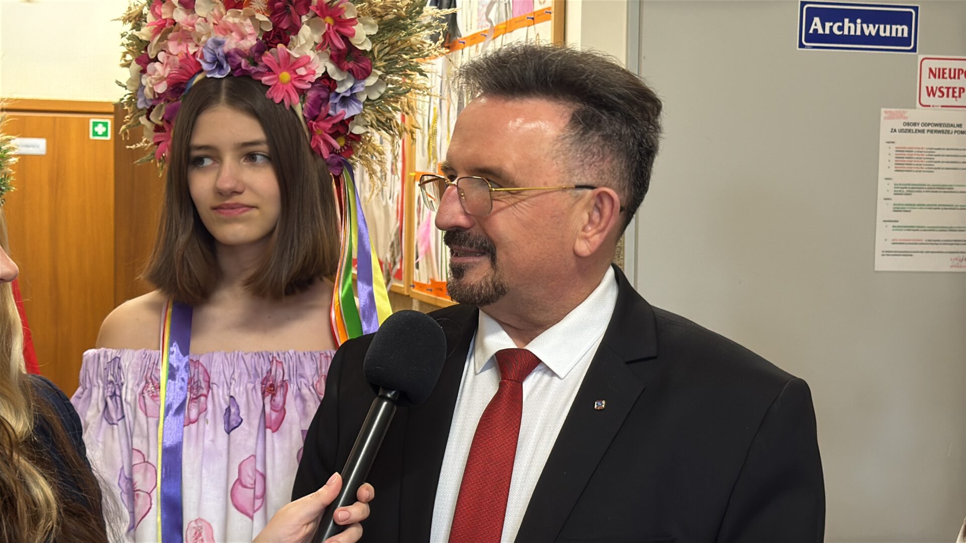 Man in a suit and red tie being interviewed with a microphone, woman in floral dress and a large flower crown stands behind him in a backstage area.
