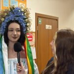 Woman wearing a large blue floral headdress with multicolored ribbons is interviewed with a handheld microphone in a school hallway.