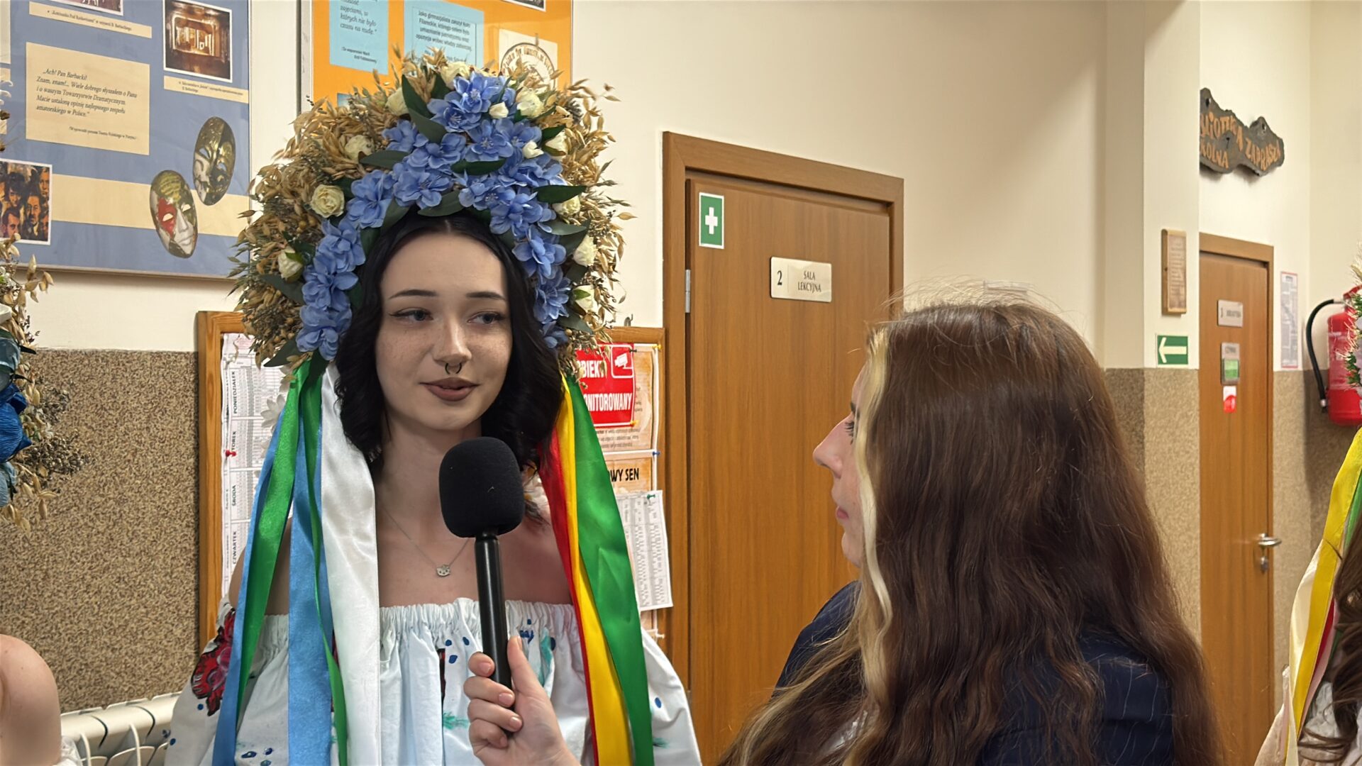 Woman wearing a large blue floral headdress with multicolored ribbons is interviewed with a handheld microphone in a school hallway.