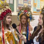 Three young women wearing elaborate floral crowns and bright ribbon accents are being interviewed by a reporter holding a microphone in a hallway.