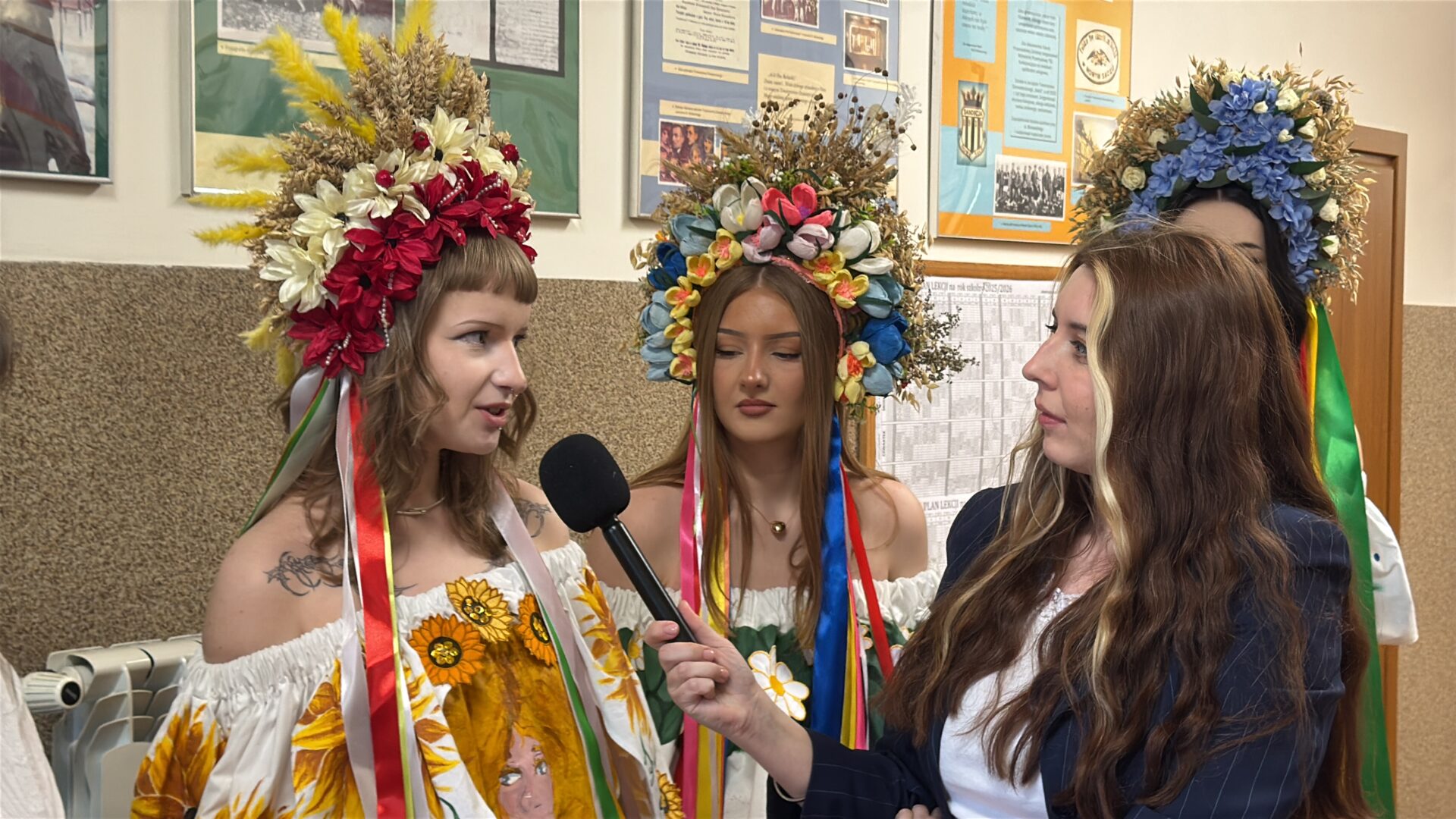 Three young women wearing elaborate floral crowns and bright ribbon accents are being interviewed by a reporter holding a microphone in a hallway.