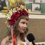 Woman with a large red-and-white flower crown speaks into a handheld microphone indoors.