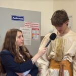 A female interviewer holds a microphone to a young man in a beige traditional costume as two women in folk outfits stand nearby in a hallway with a wreath on the left.