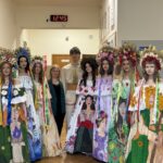 Group of students in vibrant floral-printed Ukrainian folk dresses with large flower wreaths, standing in a school hallway with a woman in a green blazer and a tall student in traditional white attire nearby.
