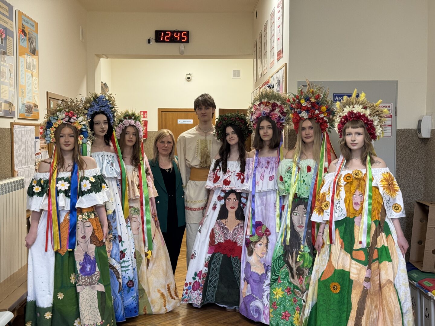 Group of students in vibrant floral-printed Ukrainian folk dresses with large flower wreaths, standing in a school hallway with a woman in a green blazer and a tall student in traditional white attire nearby.