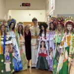Group of young women in vibrant, hand-painted folk dresses wearing large flower crowns, posing with two adults in a hallway.