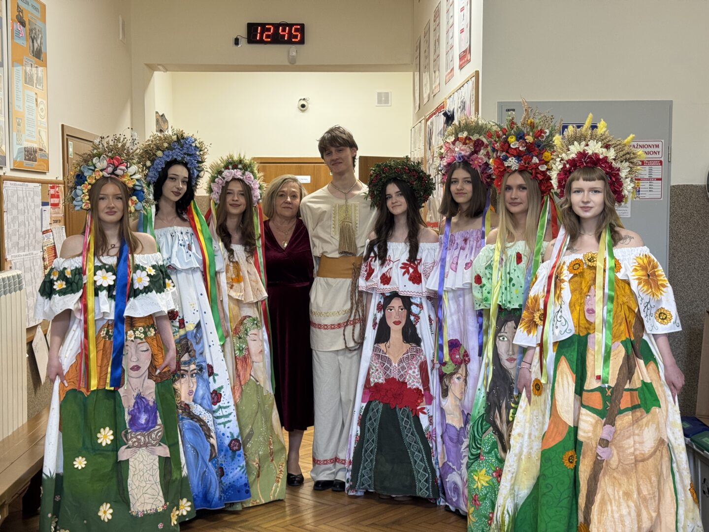 Group of young women in vibrant, hand-painted folk dresses wearing large flower crowns, posing with two adults in a hallway.