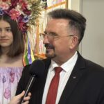 Man in suit being interviewed with a microphone, standing next to a young woman wearing a flower crown and floral dress in a hallway.
