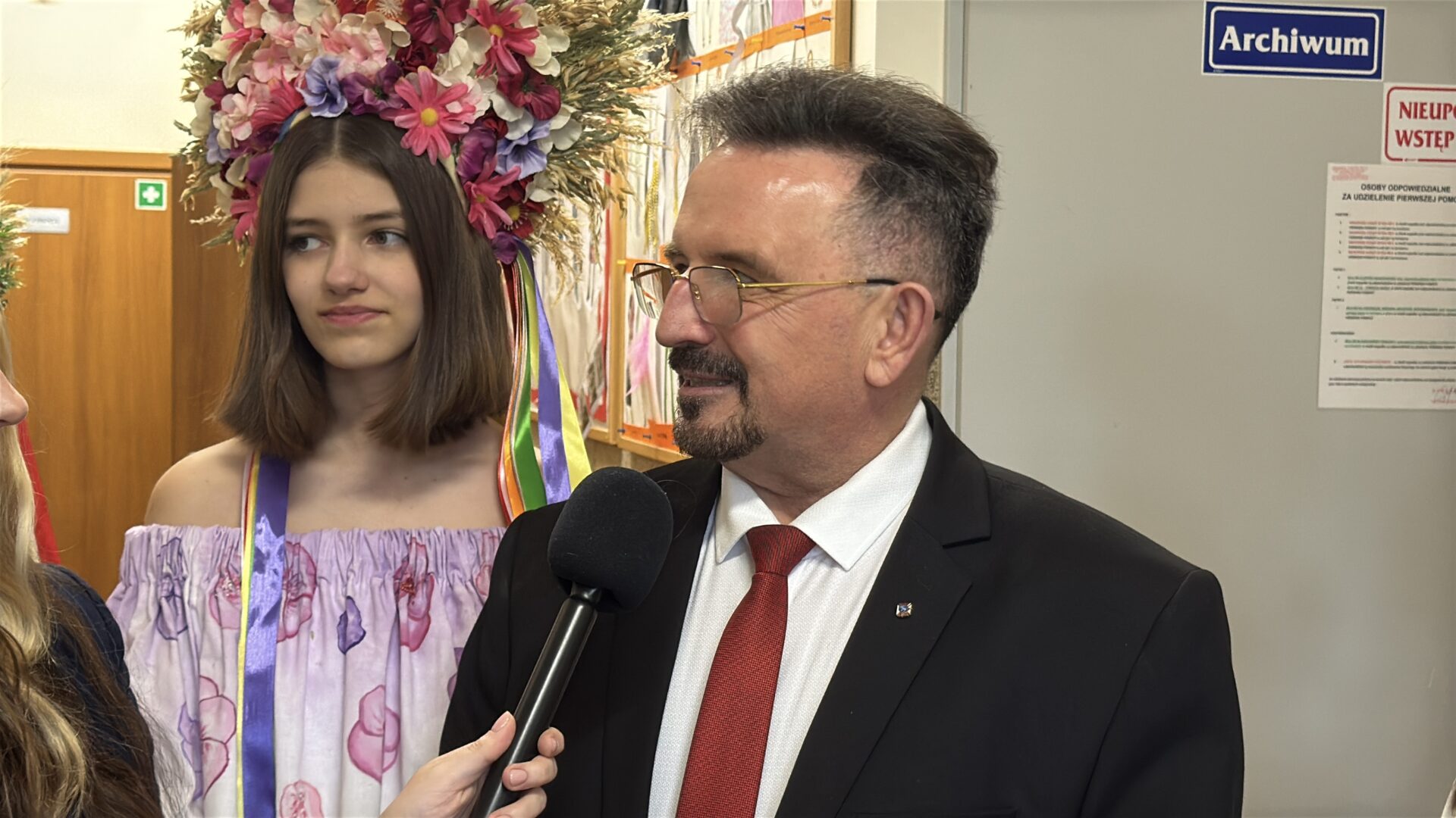 Man in suit being interviewed with a microphone, standing next to a young woman wearing a flower crown and floral dress in a hallway.