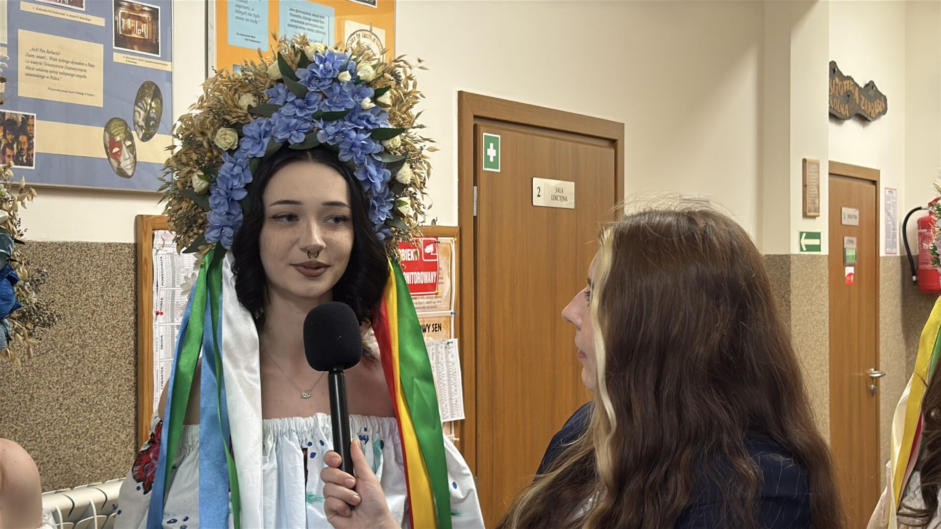 Woman in a large blue floral headpiece being interviewed with a handheld microphone in a school hallway.