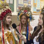 Three young women wearing large colorful flower crowns and festive dresses are being interviewed indoors, with a reporter holding a microphone to the left speaker.