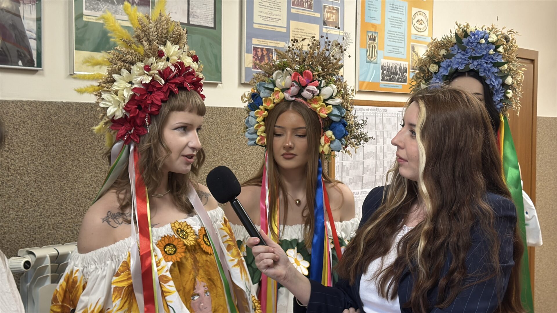 Three young women wearing large colorful flower crowns and festive dresses are being interviewed indoors, with a reporter holding a microphone to the left speaker.