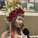 Woman wearing a large, colorful flower crown is being interviewed with a handheld microphone nearby.