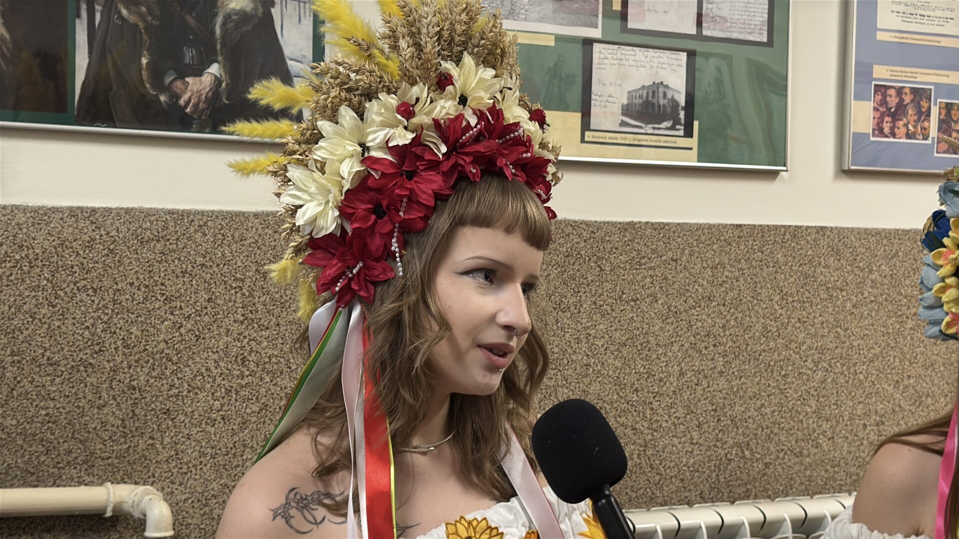 Woman wearing a large, colorful flower crown is being interviewed with a handheld microphone nearby.
