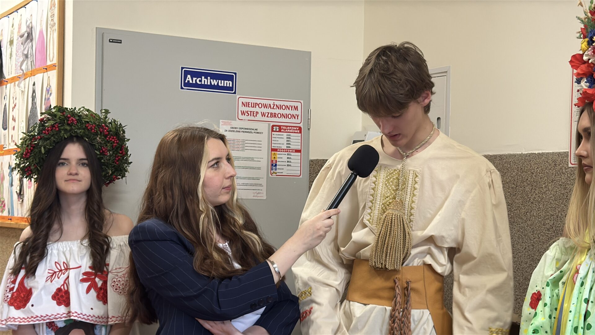 A woman interviews a young man in traditional beige costume with tassels, holding a microphone in a school hallway.
