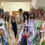 Group of young women in bright floral folk dresses and large flower crowns, posing with two adults in a hallway.