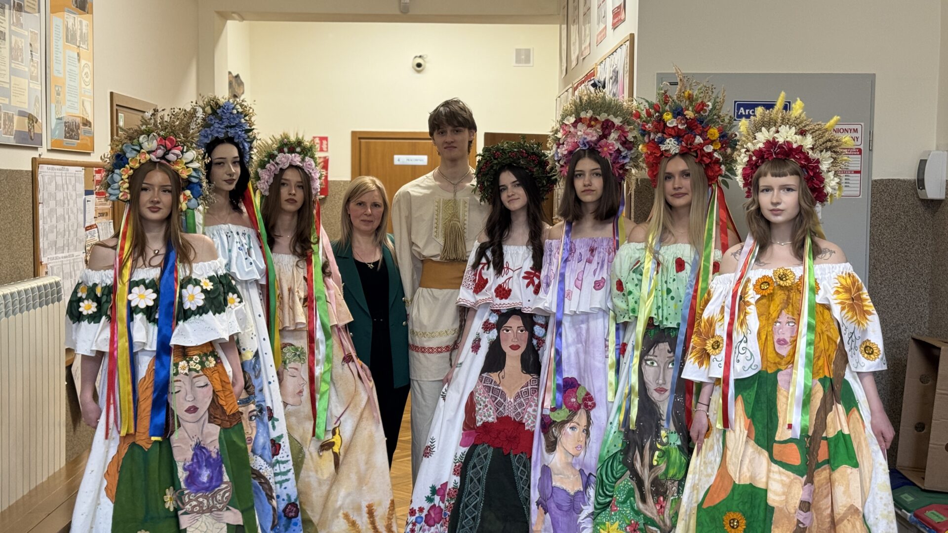 Group of young women in bright floral folk dresses and large flower crowns, posing with two adults in a hallway.