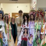 Group of young people in colorful, flower-adorned Ukrainian‑style dresses with large floral crowns posing in a school hallway with bulletin boards behind them.