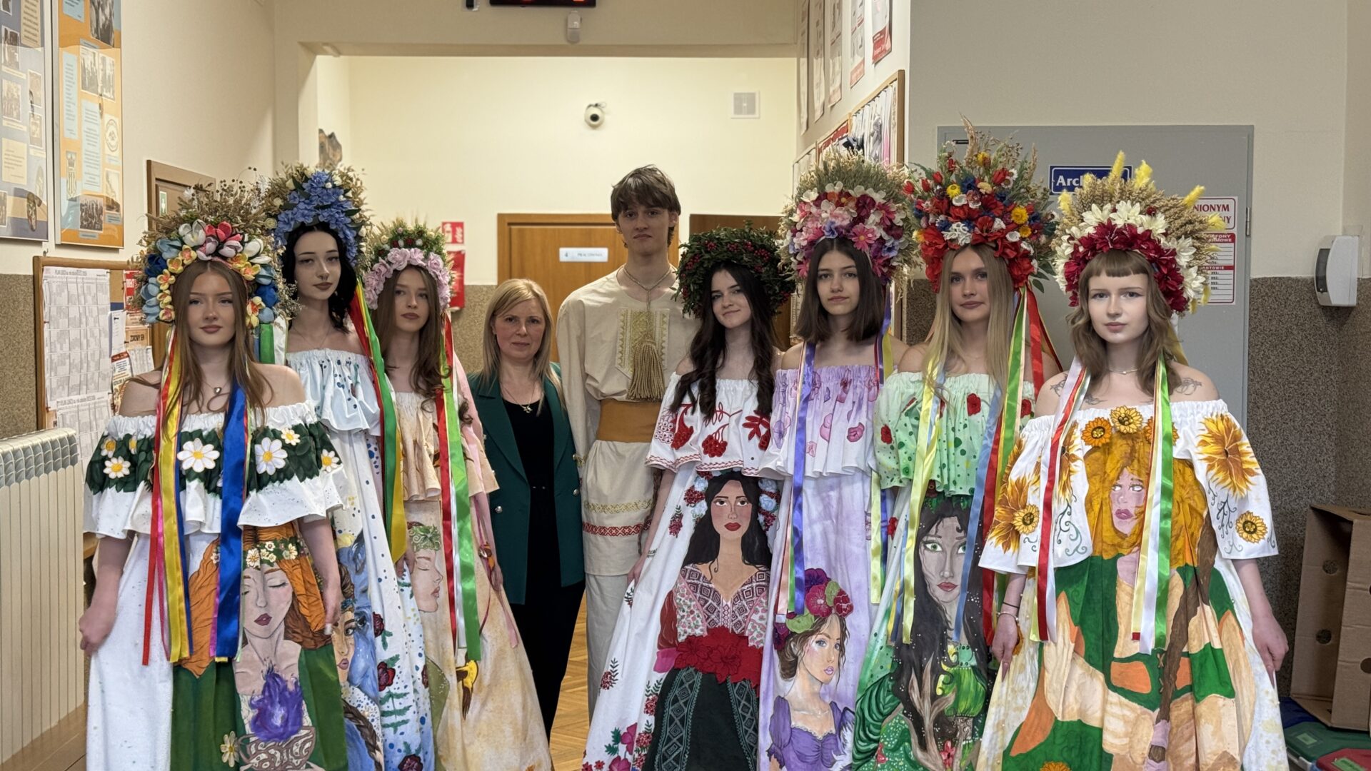 Group of young people in colorful, flower-adorned Ukrainian‑style dresses with large floral crowns posing in a school hallway with bulletin boards behind them.