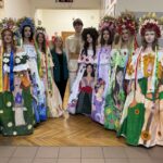 Group of people in colorful traditional dresses with floral wreaths posing in a school hallway (performers in folk costumes).