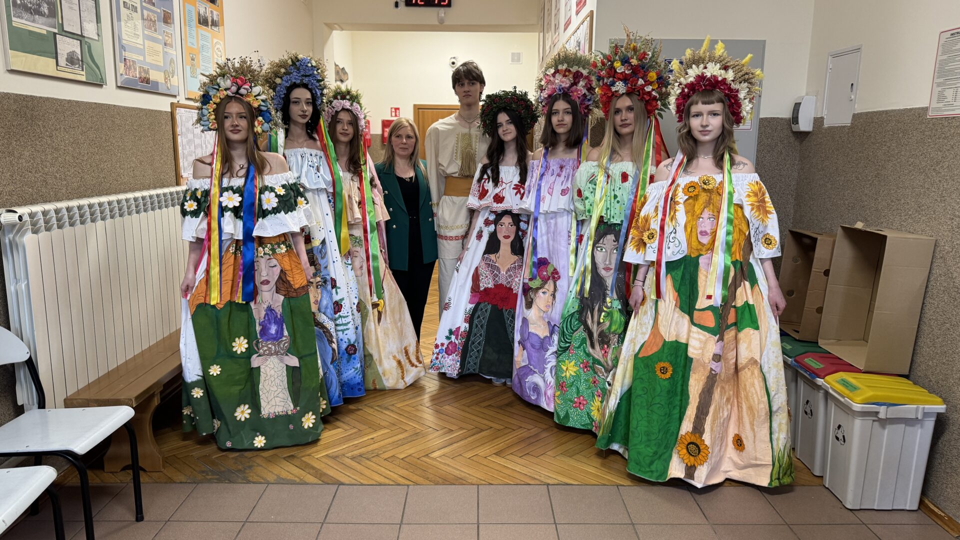 Group of people in colorful traditional dresses with floral wreaths posing in a school hallway (performers in folk costumes).
