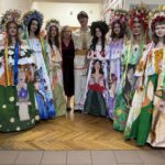 Group of eight young people in bright, flower-themed dresses and flower crowns posing in a school hallway with two adults behind them.