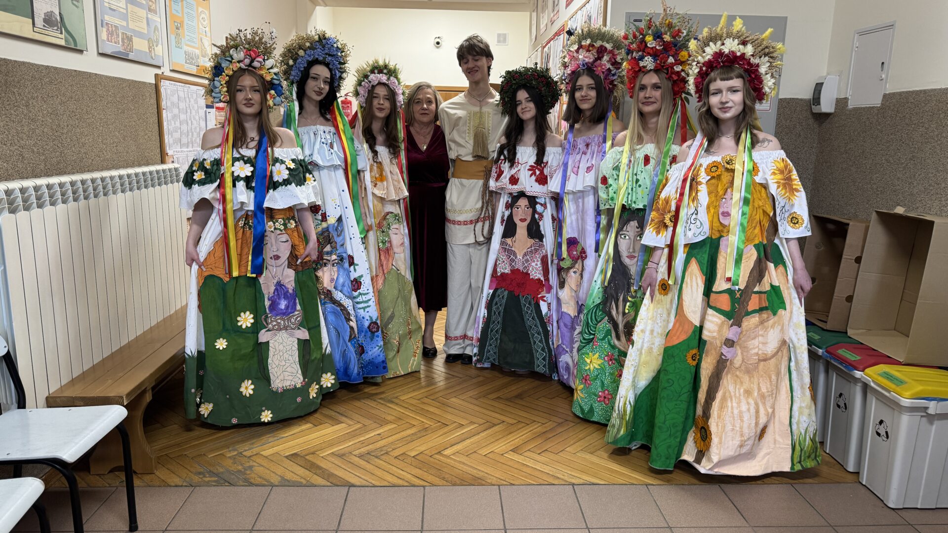 Group of eight young people in bright, flower-themed dresses and flower crowns posing in a school hallway with two adults behind them.
