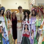 Group of eight people in a hallway wearing large floral wreaths and illustrated dresses, posing for a photo with two adults in the center.