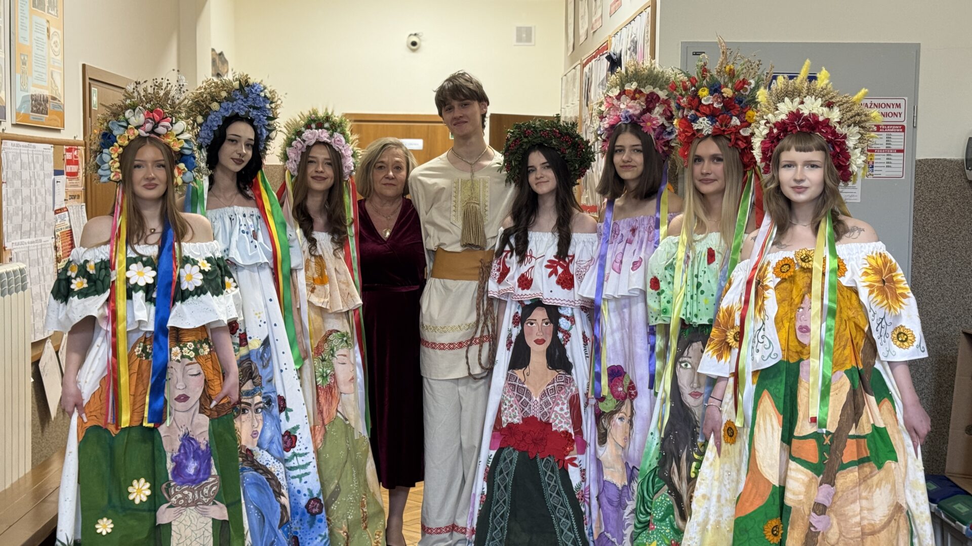 Group of eight people in a hallway wearing large floral wreaths and illustrated dresses, posing for a photo with two adults in the center.