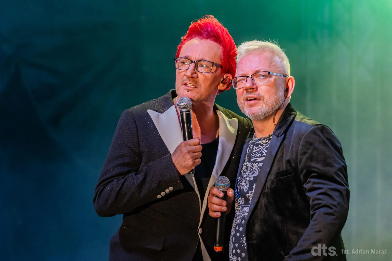 Two male performers sing into handheld microphones on stage, one with bright red hair and glasses, leaning close together under stage lights.
