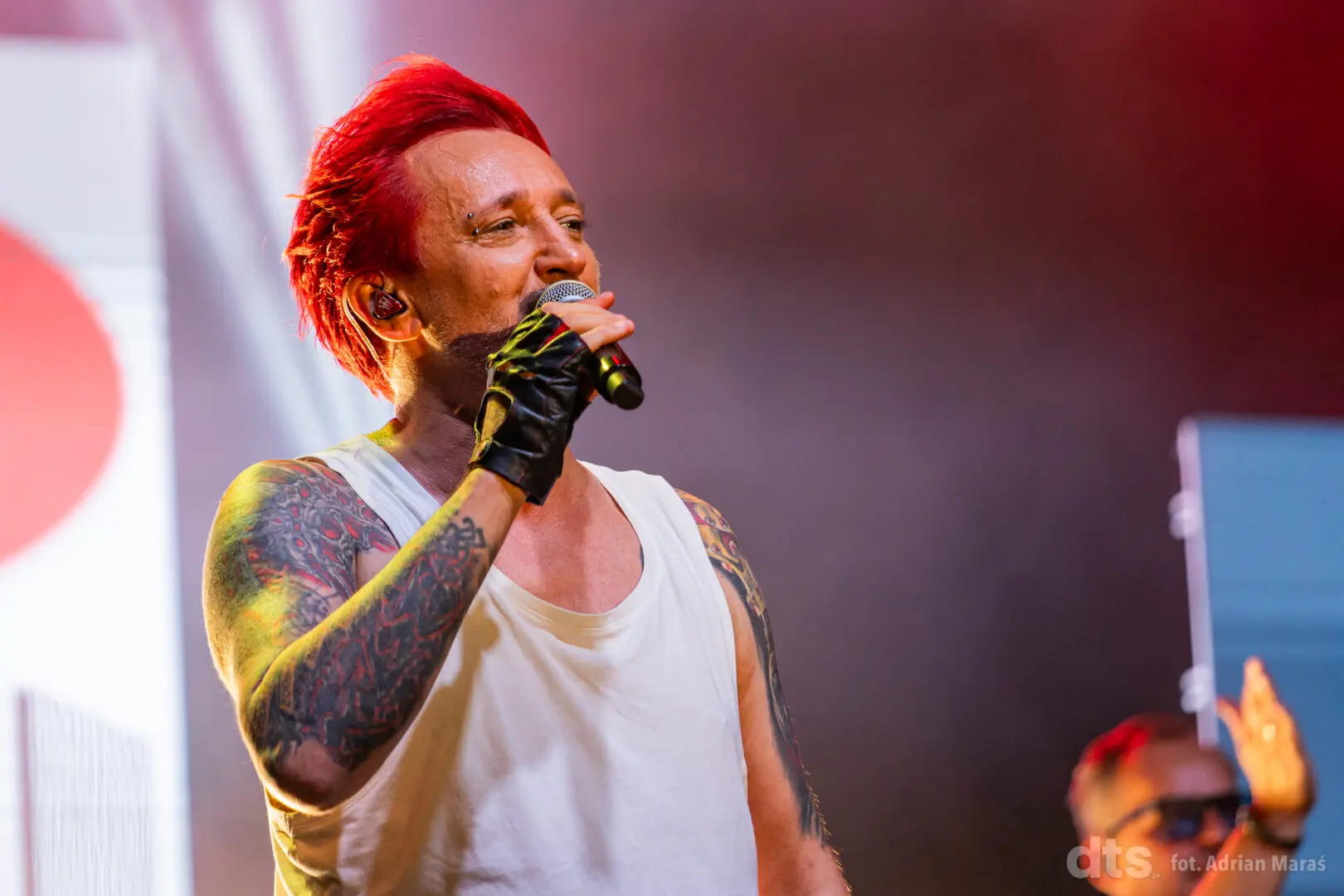 Male singer with bright red hair, white sleeveless shirt, and full-arm tattoos performing on stage while holding a microphone