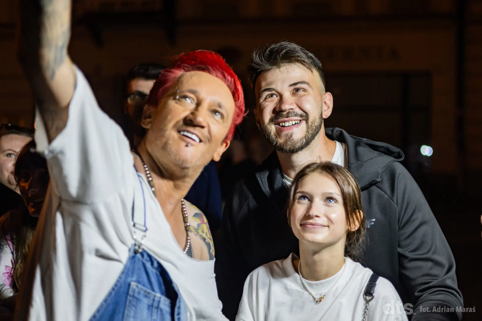 Three friends posing for a selfie at night, with a man raising his arm and a smiling couple close together in front.