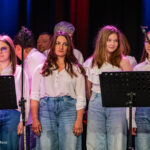 Group of female singers on stage in white tops and blue jeans, standing behind music stands with microphones nearby, looking toward the audience.