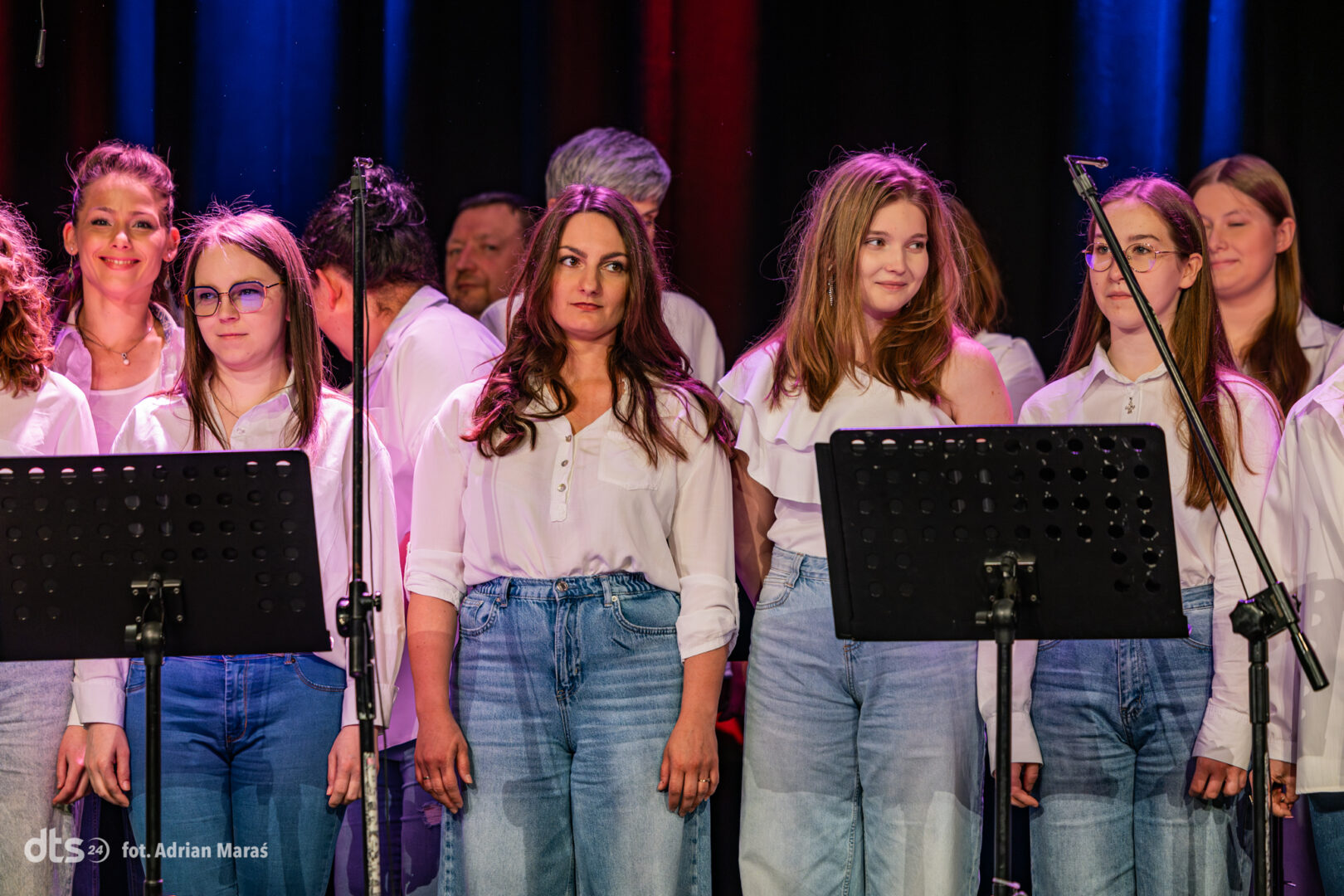 Group of female singers on stage in white tops and blue jeans, standing behind music stands with microphones nearby, looking toward the audience.