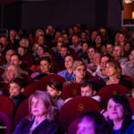 Audience seated in a dark theater, watching a performance; red upholstered seats with numbered labels crowding the venue.