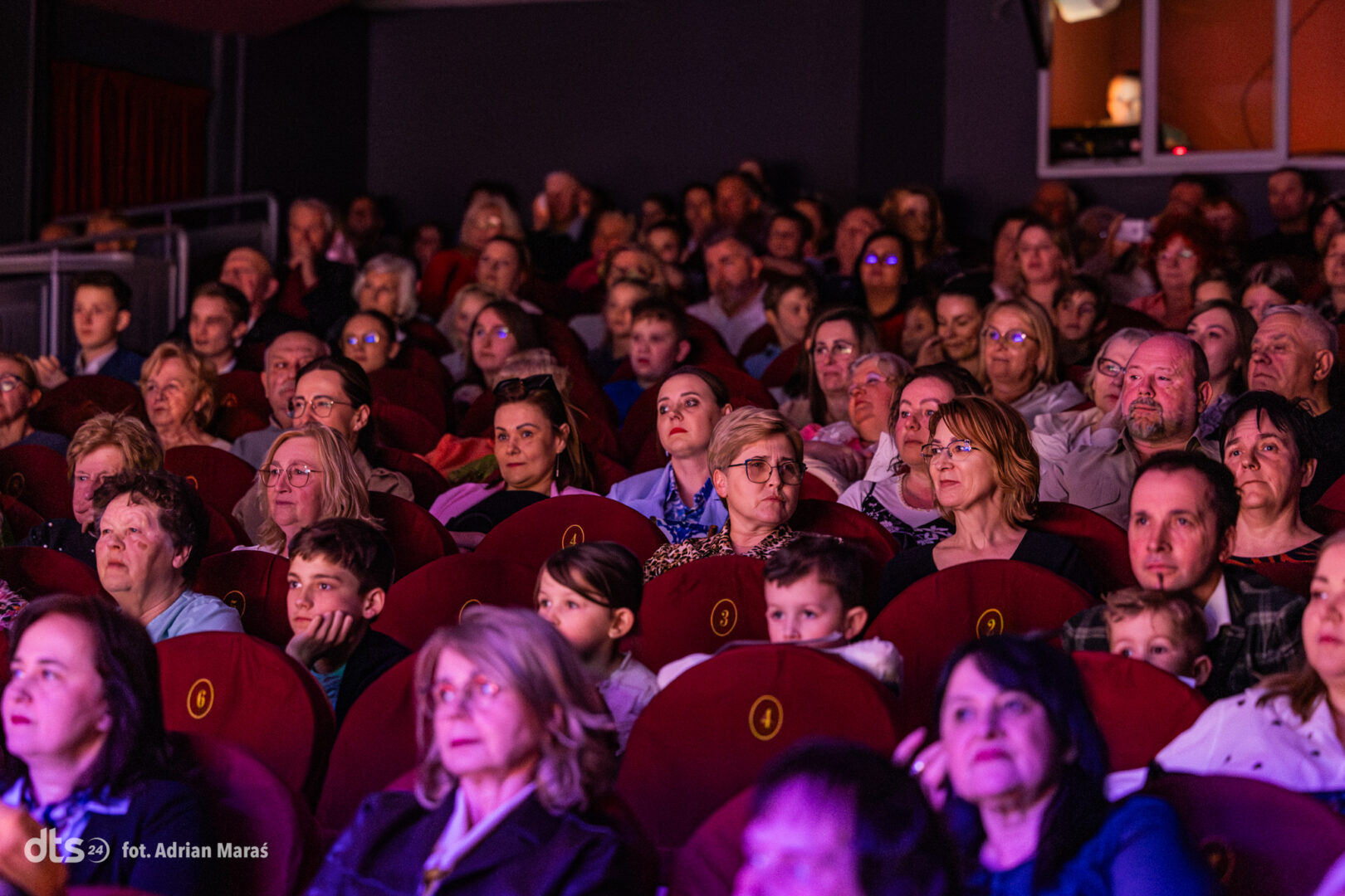 Audience seated in a dark theater, watching a performance; red upholstered seats with numbered labels crowding the venue.