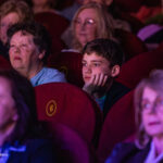 Audience members watching a performance; a young boy rests his chin on his hand, looking thoughtful amid red theater seats.