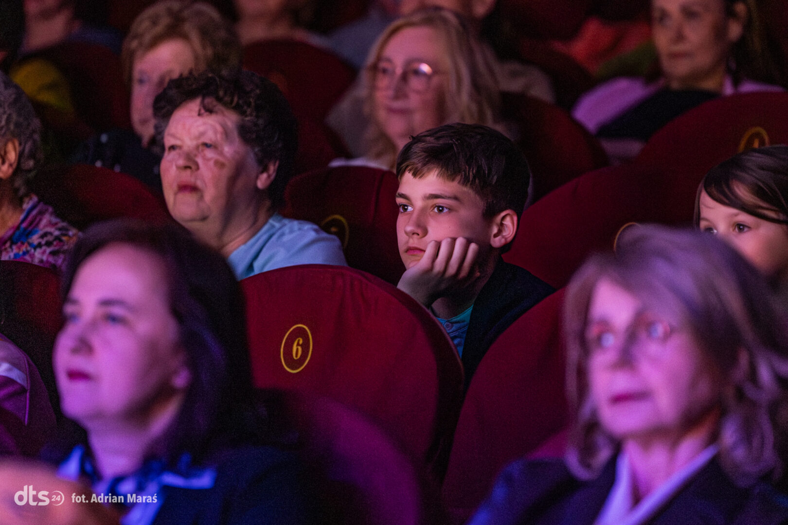 Audience members watching a performance; a young boy rests his chin on his hand, looking thoughtful amid red theater seats.