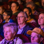 Audience seated in a theater, faces lit by stage lighting as they watch a performance.