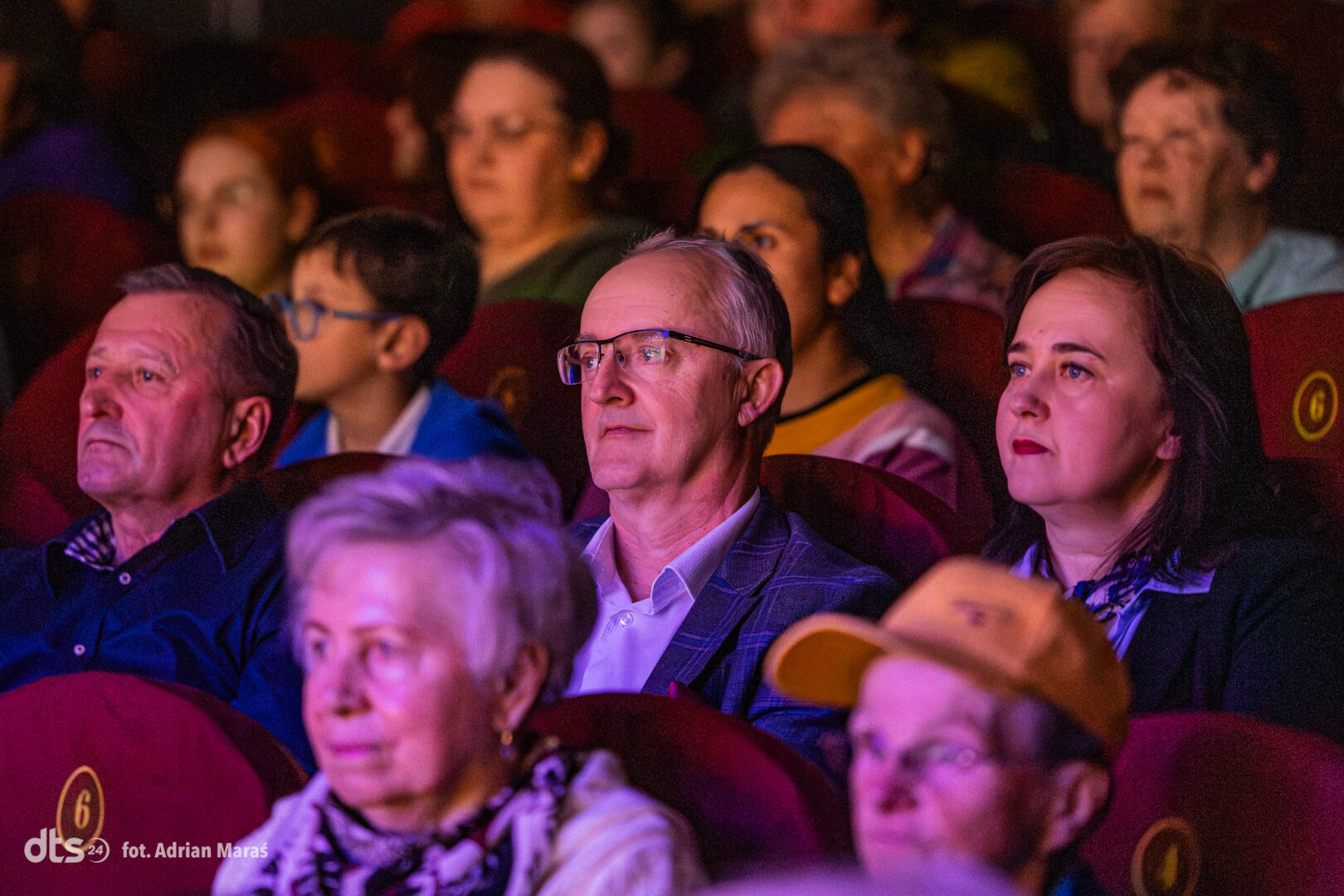 Audience seated in a theater, faces lit by stage lighting as they watch a performance.
