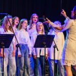 Chorus of young women singing on stage under blue lighting, led by a conductor in a white lace dress.