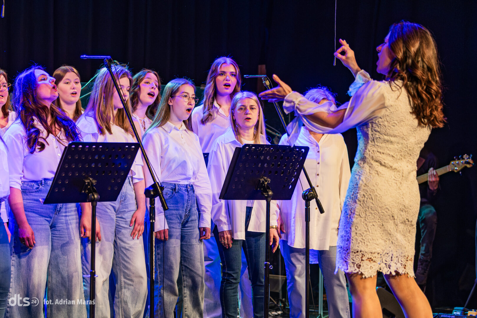 Chorus of young women singing on stage under blue lighting, led by a conductor in a white lace dress.