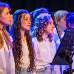 A group of young singers in white shirts perform on stage with a music stand and microphone.