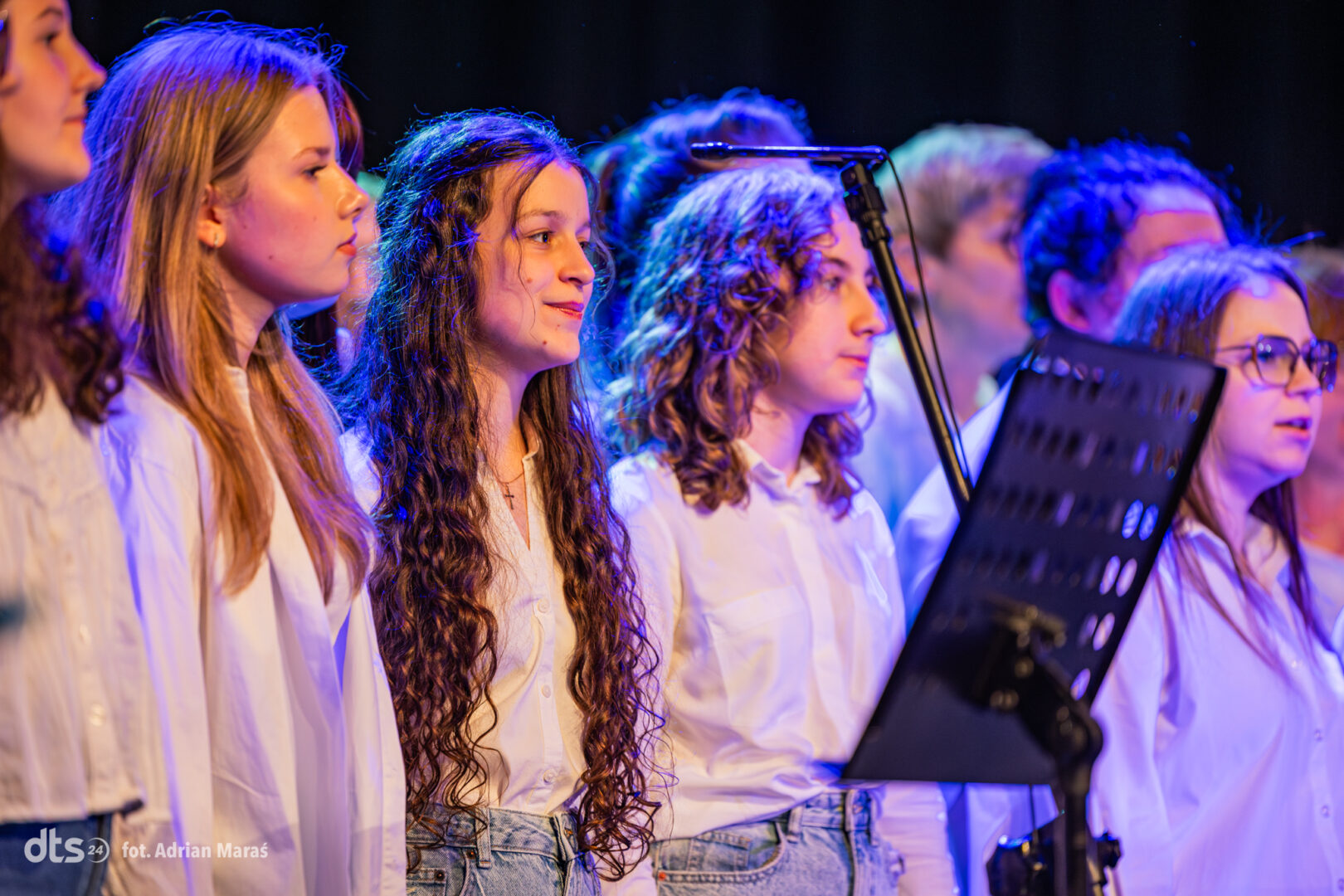 A group of young singers in white shirts perform on stage with a music stand and microphone.