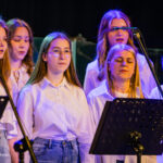 Group of young women in white shirts singing on stage with music stands and microphones under blue stage lighting, as a choir performance.