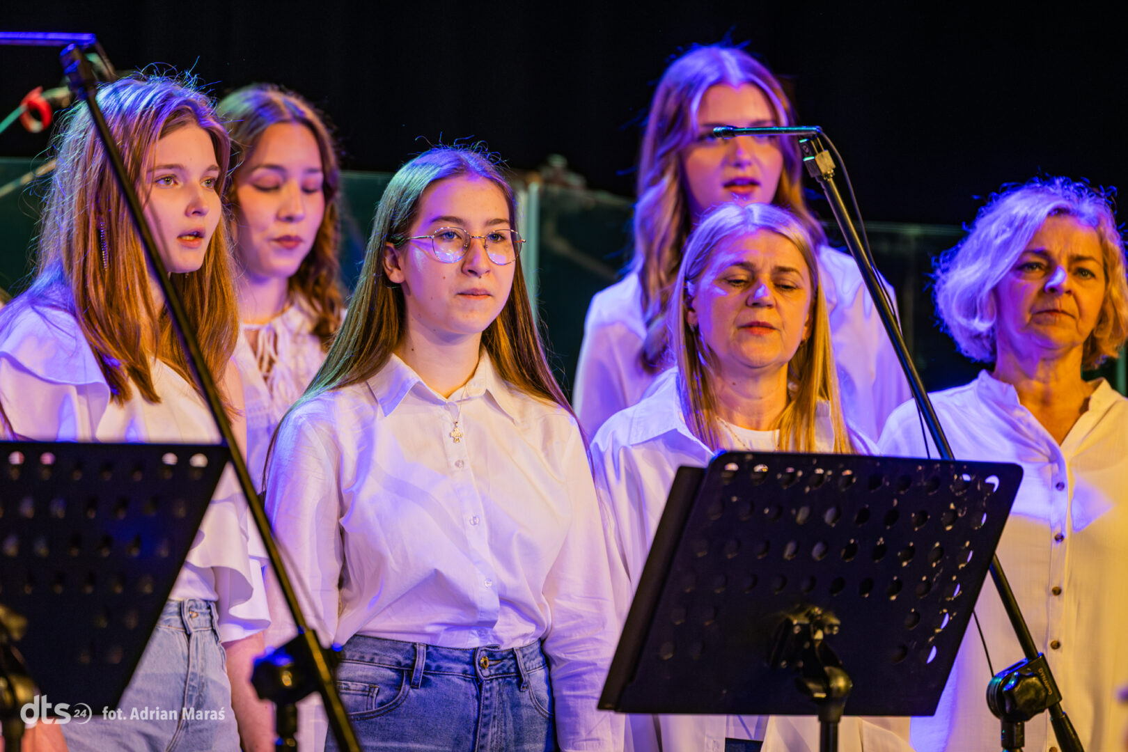 Group of young women in white shirts singing on stage with music stands and microphones under blue stage lighting, as a choir performance.
