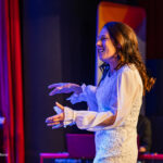 Female singer in a white lace dress on stage, smiling with hands raised as a guitarist watches in the background under colorful stage lighting.