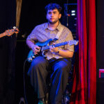 Musician seated on stage playing a blue electric guitar, with a pedalboard at his feet and red stage curtains beside him.